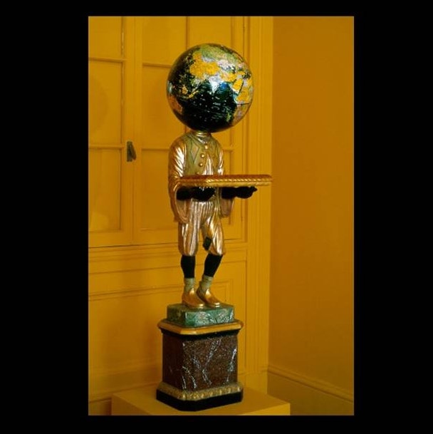 Art work in the figure of a man with a globe of the world for its head, carrying a pillow, evoking posture of a servant.