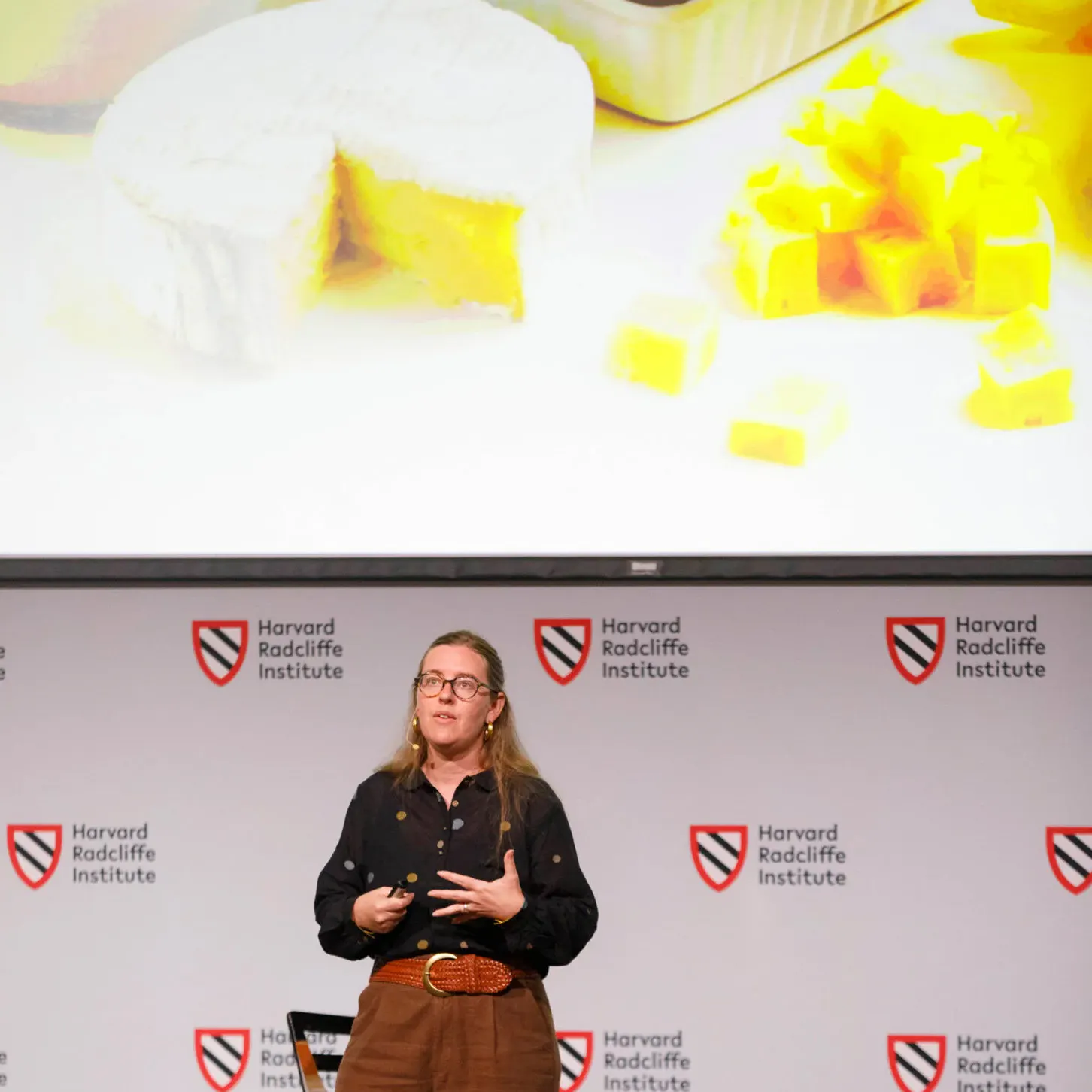 A woman stands on a stage, talking, below a giant projection of different cheeses.