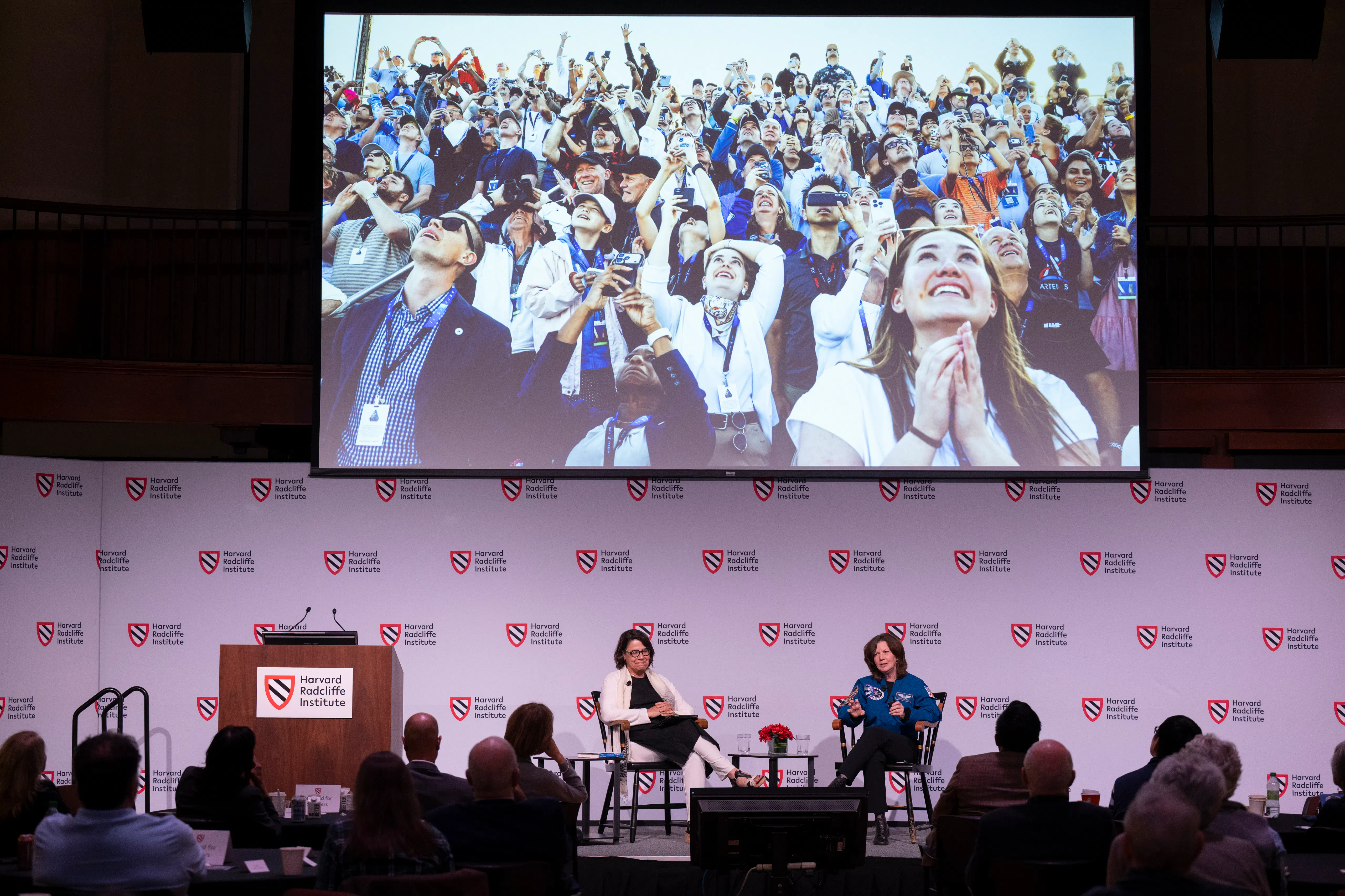 Two women sitting in chairs chat onstage while a photo of a large group of joyful people looking up as they snap photos with their smartphones looms large on the screen overhead.