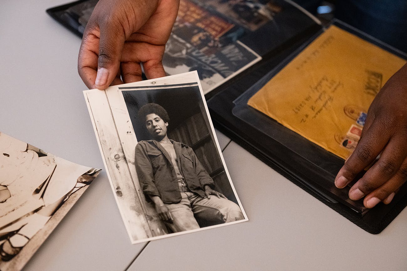 Close up of hands holding a vintage black-and-white photograph of a softly smiling, casually dressed young Black woman with a short afro leaning against a doorframe.