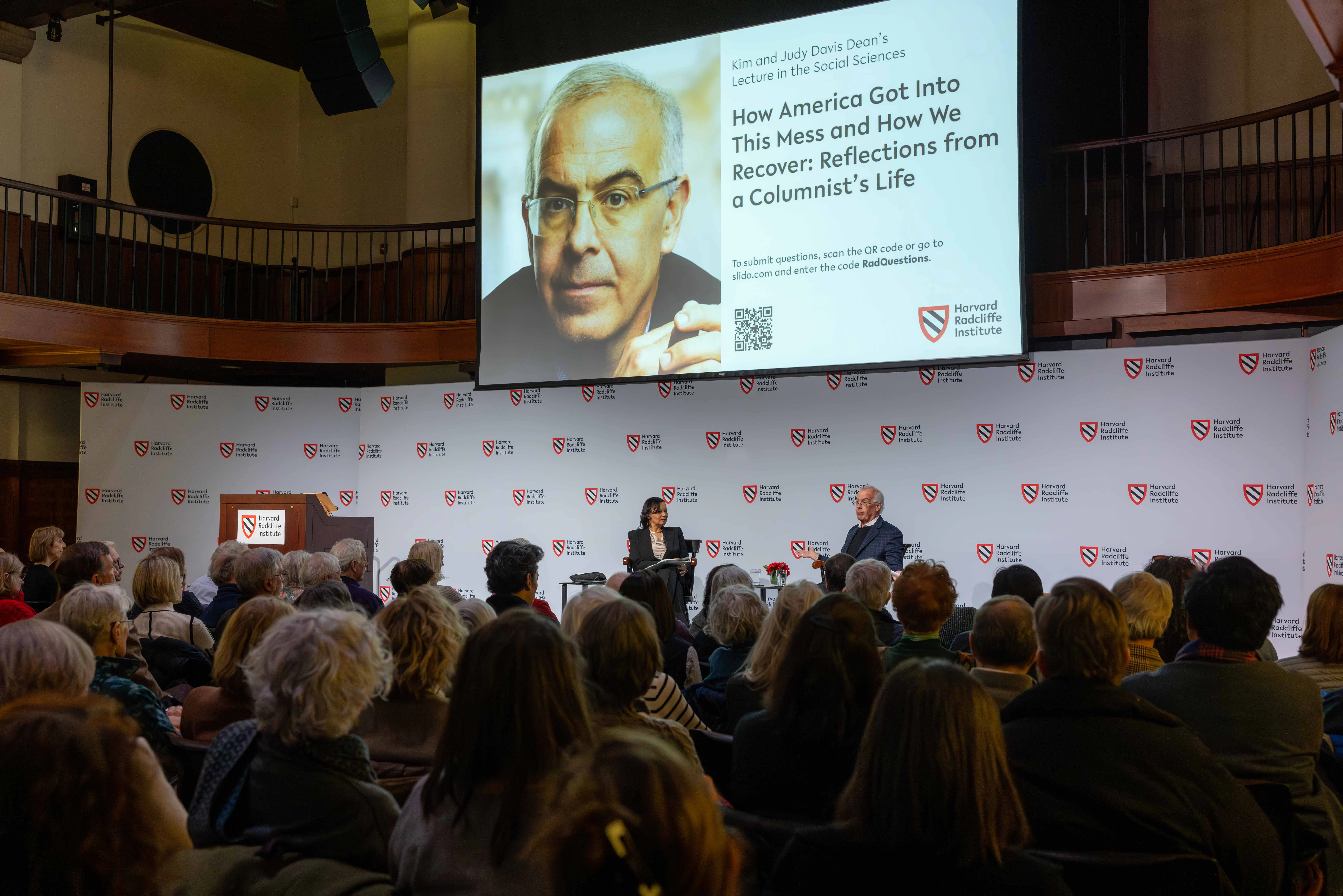 Packed event auditorium with man and woman speaking on stage