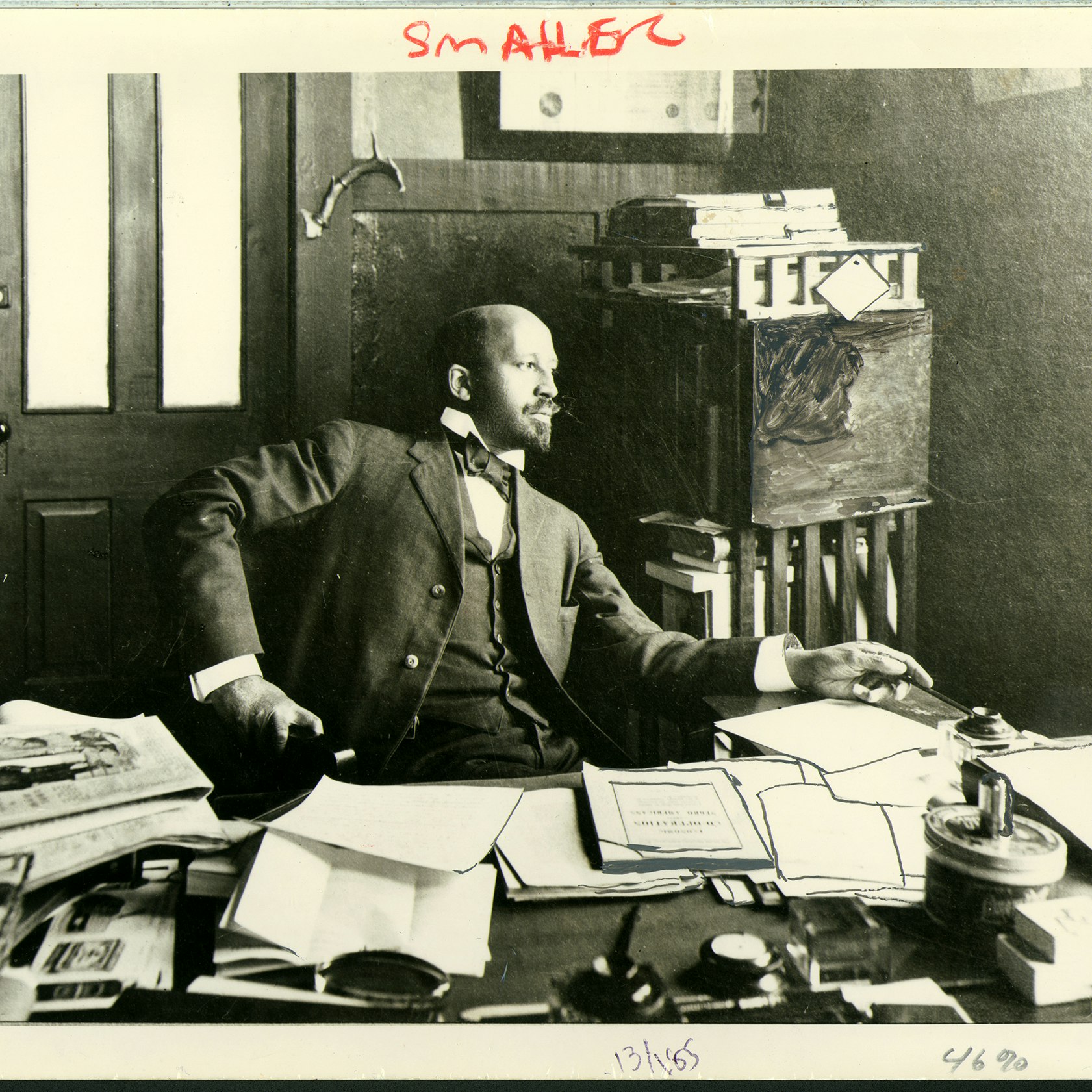 W. E. B. Du Bois seated at his desk in his office at Atlanta University. There are many papers on the desk and Du Bois is leaning back and looking off into the distance.