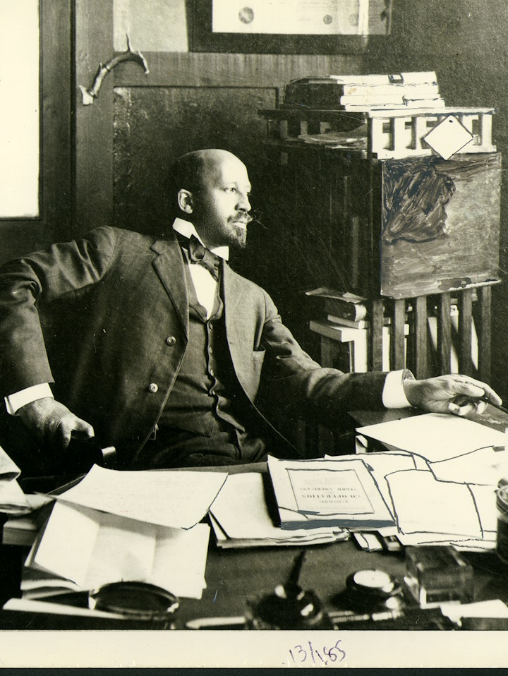 W. E. B. Du Bois seated at his desk in his office at Atlanta University. There are many papers on the desk and Du Bois is leaning back and looking off into the distance.