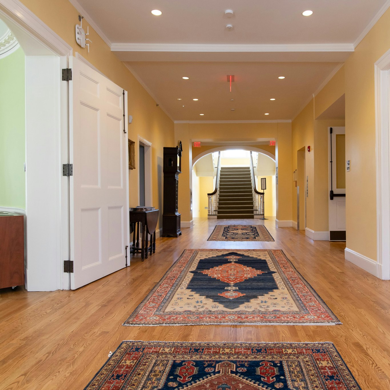 Lobby with wood floors and three printed rugs. At the end of the lobby there is a dark brown staircase leading upstairs.