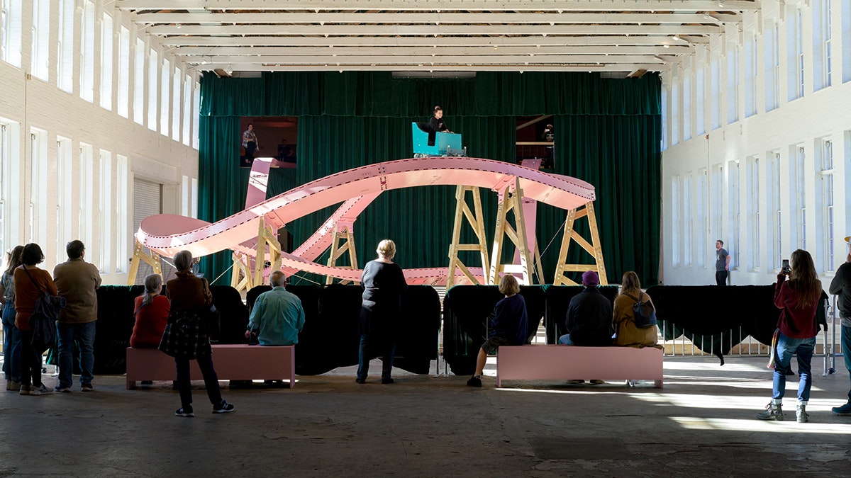 Spectators watch as someone rides a small-scale pink roller coaster inside a large warehouse building