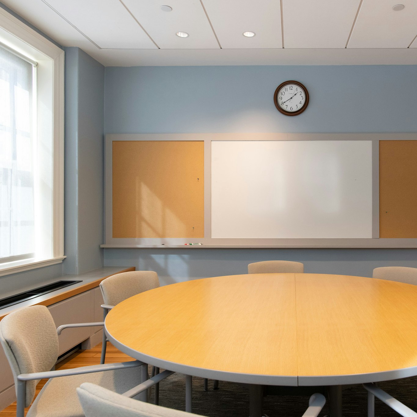 Circular conference room table, seating for 7 people. To the right is an entrance of two white double doors, and to the left are some windows letting in natural light. In the back center of the room is a whiteboard and a clock mounted directly above it.