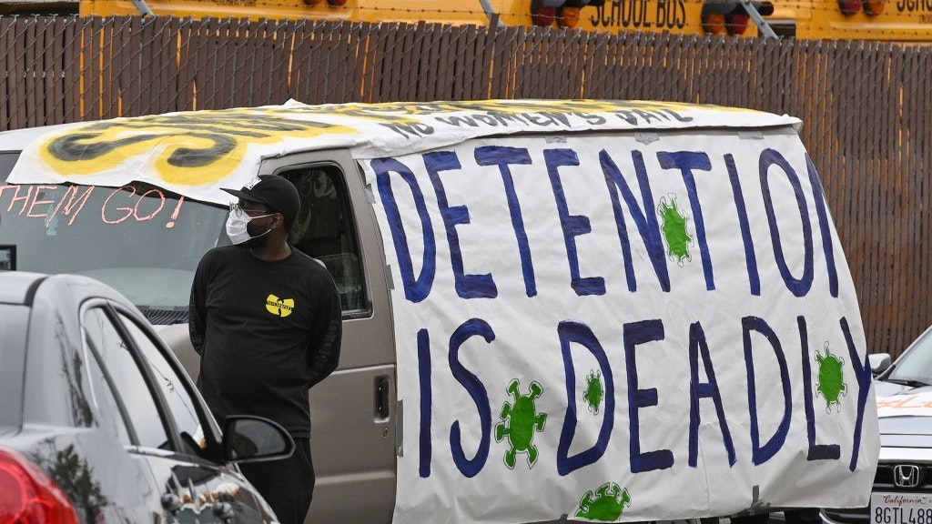 A man wearing a face covering and a Wu-Tang sweatshirt stands in front of a van papered over with a protest sign that reads "DETENTION IS DEADLY."
