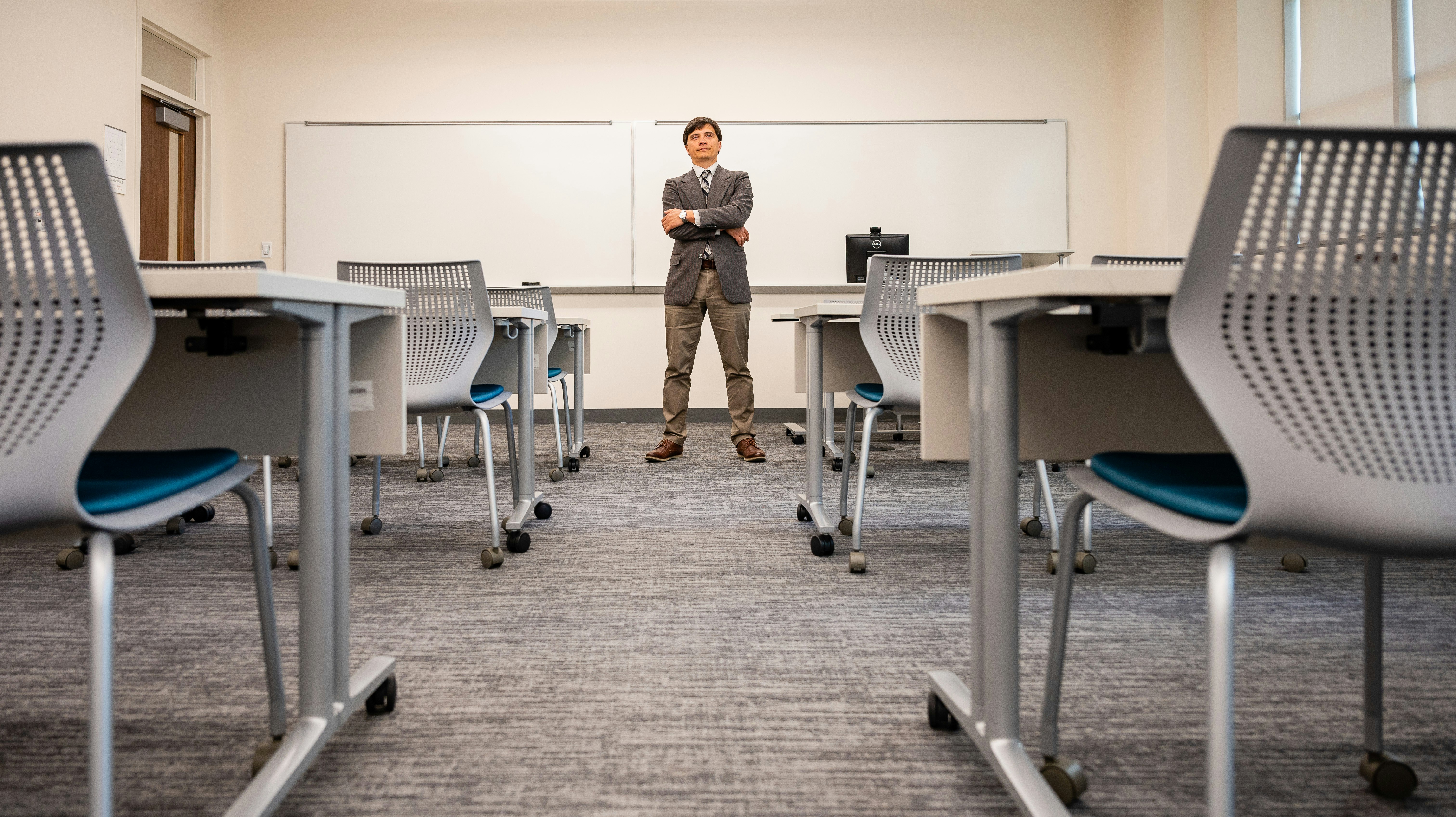 A man stands, arms crossed, in front of a classroom whiteboard. In the foreground, empty chairs.