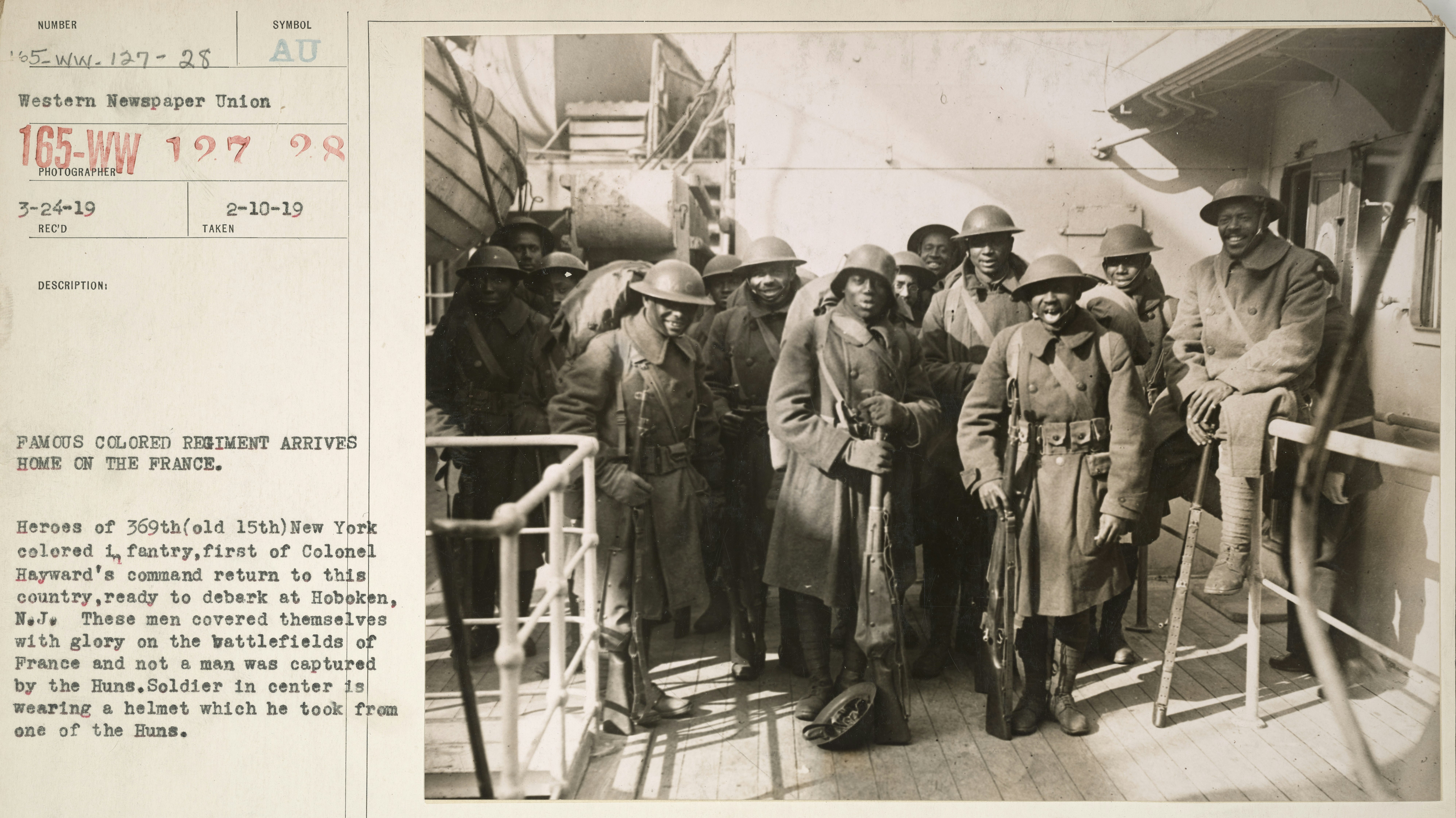 A historical photo shows a group of Black US soldiers. The original caption reads: Famous Colored Regiment Arrives Home on the France. Heroes of the 369th (old 15th) New York colored infantry, first of Colonel Hayward's command return to this country, ready to debark at Hoboken, N.J. These men covered themselves with glory on the battlefields of France and not a man was captured by the Huns. Soldier in center is wearing a helmet which he took from one of the Huns.