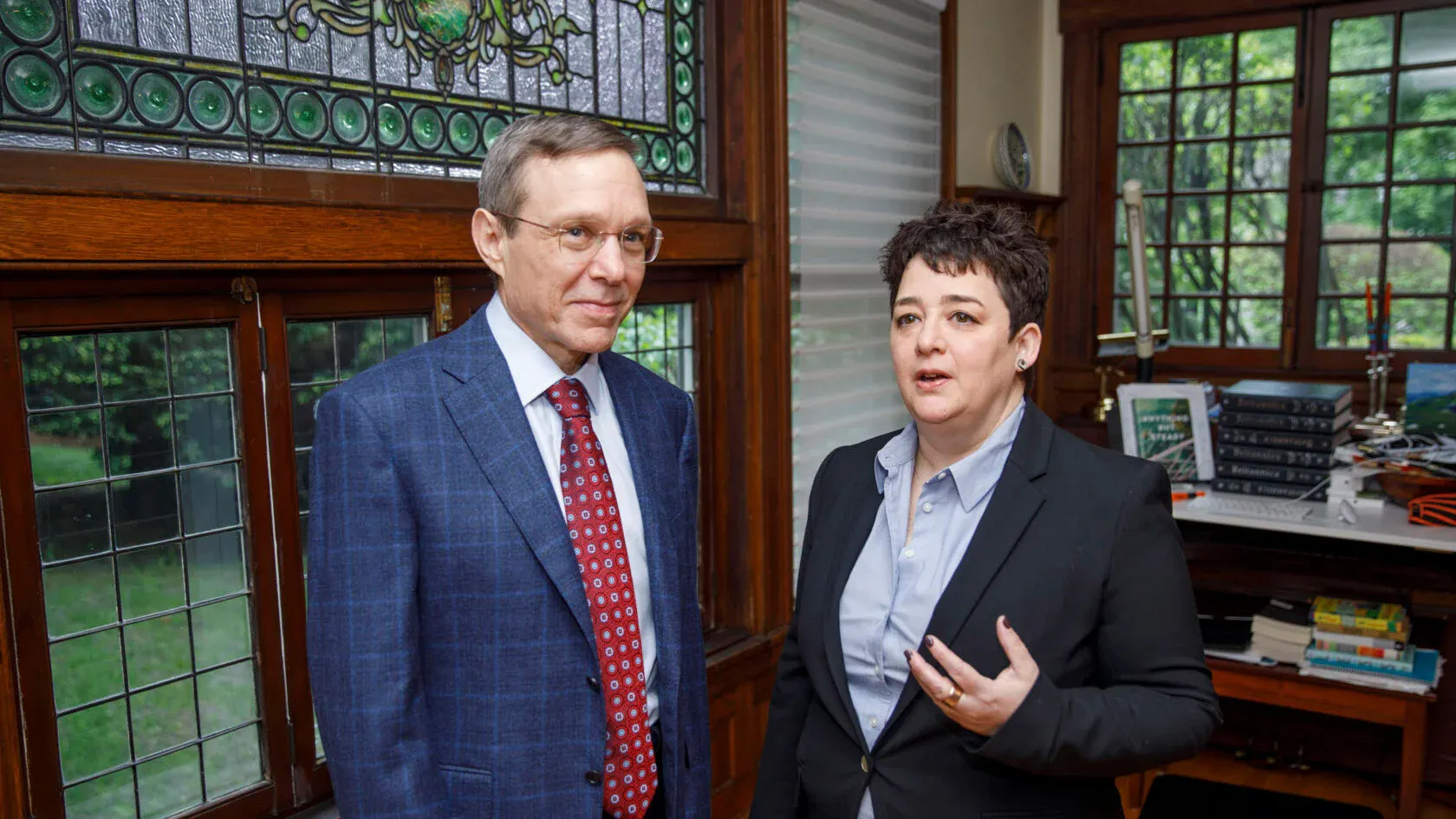 A man and a woman, both in dar suit jackets, stand indoors in front of dramatic wood-framed vintage windows, some stained glass.