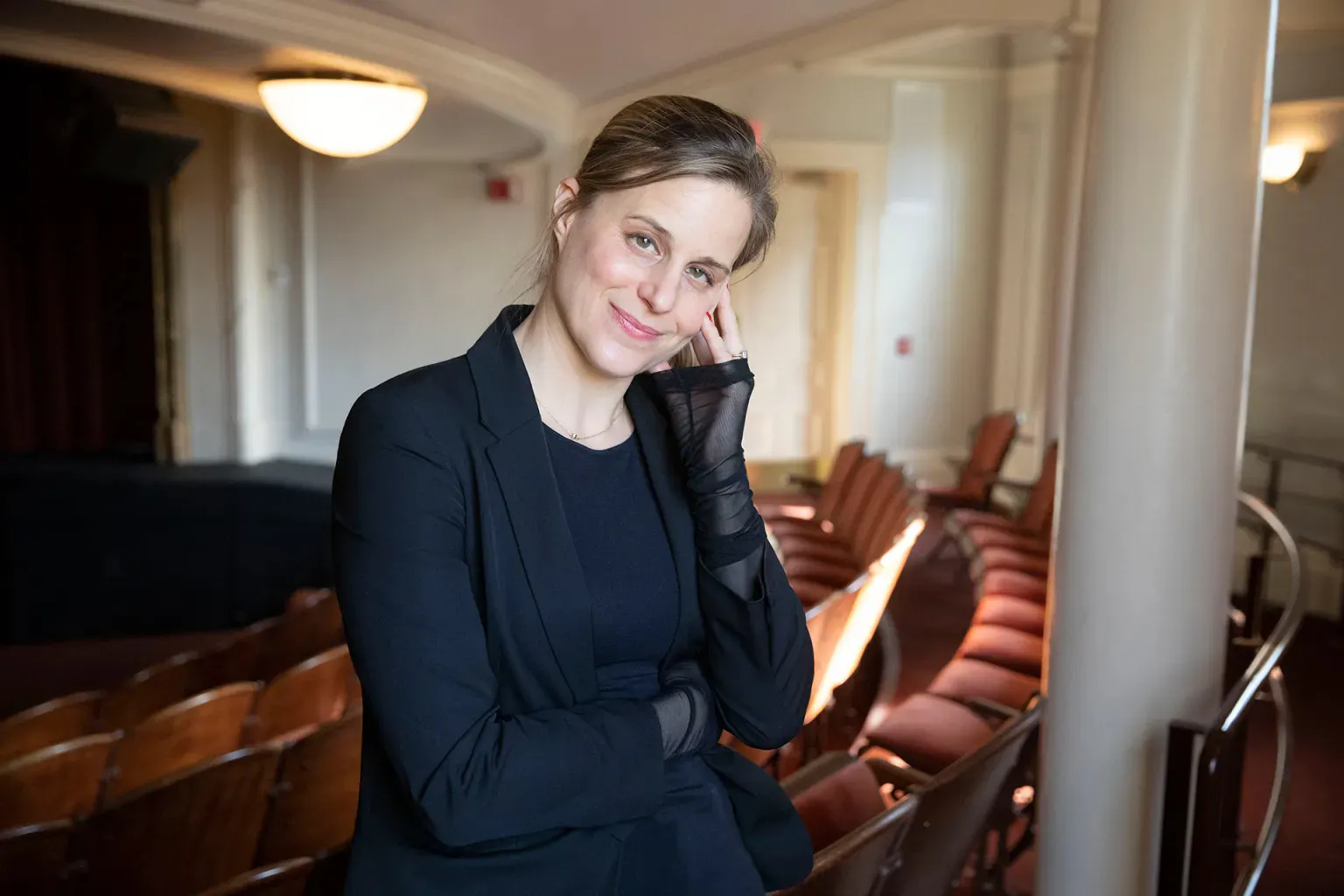A woman smiles gently, one arm crossed, the other held up to her cheek, as she stands in a theater.