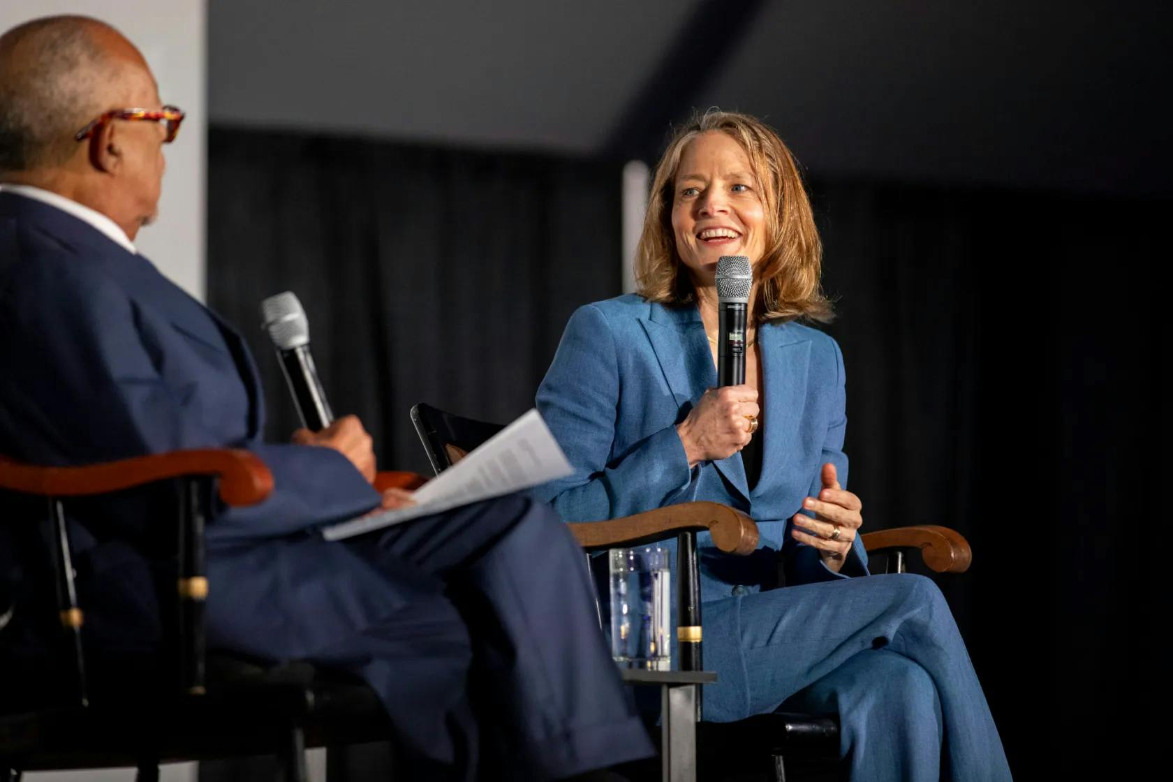 A man and woman seated in captain's chairs on a stage holding microphones. The woman is speaking.