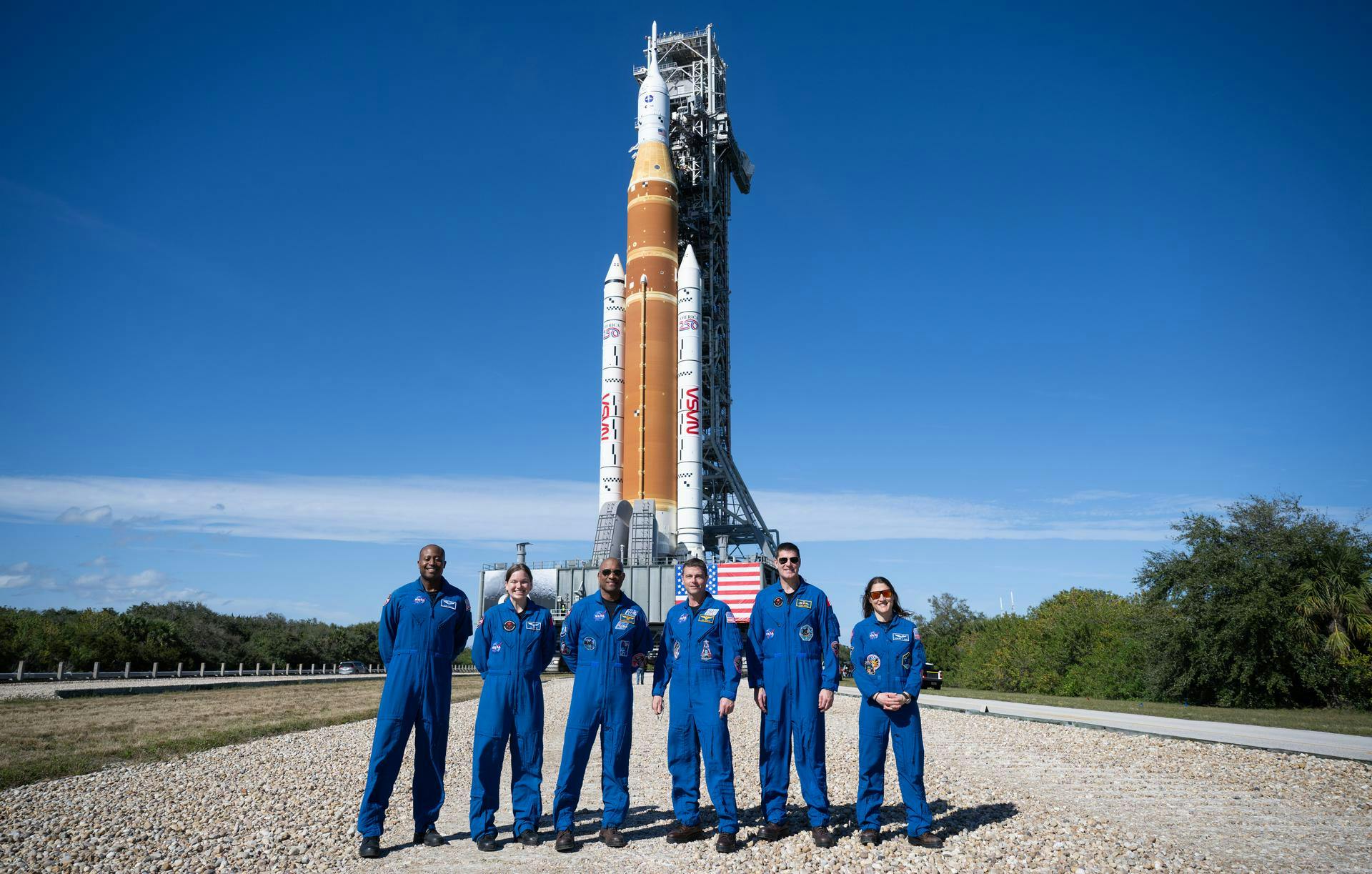Six smiling people of varying identities, all in identical blue jumpsuits, pose outdoors in a line. In the distance behind them, a rocket is poised for launch.