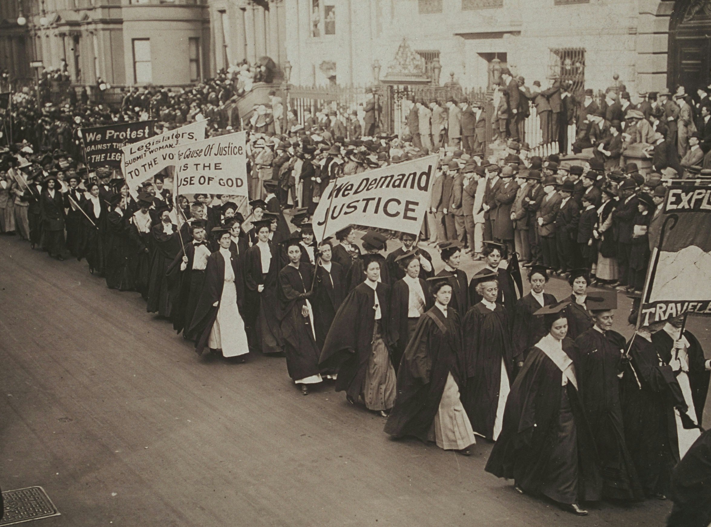 Women in academic dress march in a suffrage parade in New York City. They carry banners that read "We Demand Justice," "The Cause of Justice is the Cause of God," "Legislators, Submit Woman [cut off] To The Vote."