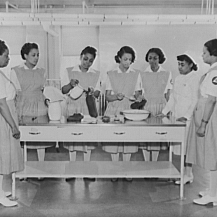 A black and white photo of six volunteer women nurse aides of color receiving training from a woman staff nurse of color.